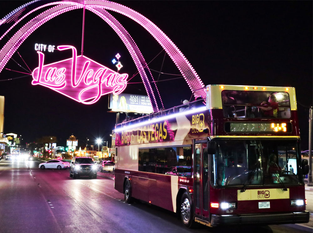 Big Bus in Las Vegas at night, passing under a neon sign 